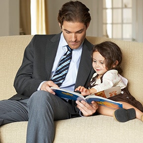 Father in suit and tie reading to his young daughter on couch