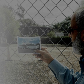 Old man in sun glasses holding photo of building behind the fence